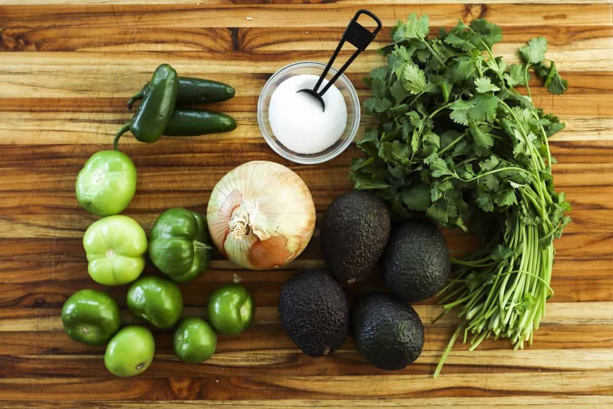 Fresh vegetables, tomatillos, avocados, onion, jalapeno, cilantro sit on a wooden cutting board.