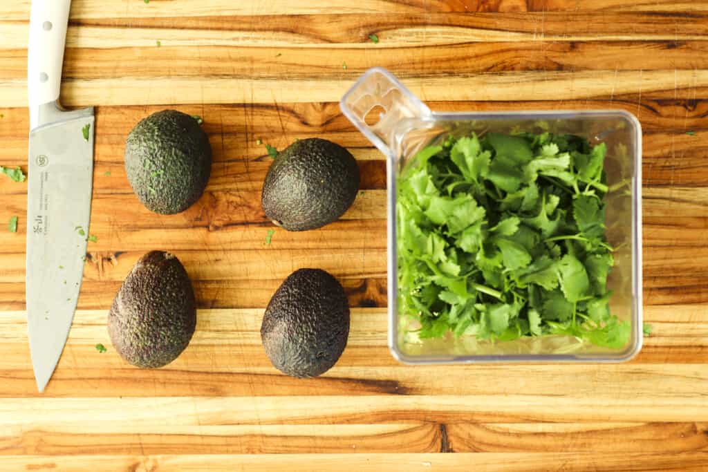 Four avocados sit next to a blender filled with chopped cilantro.