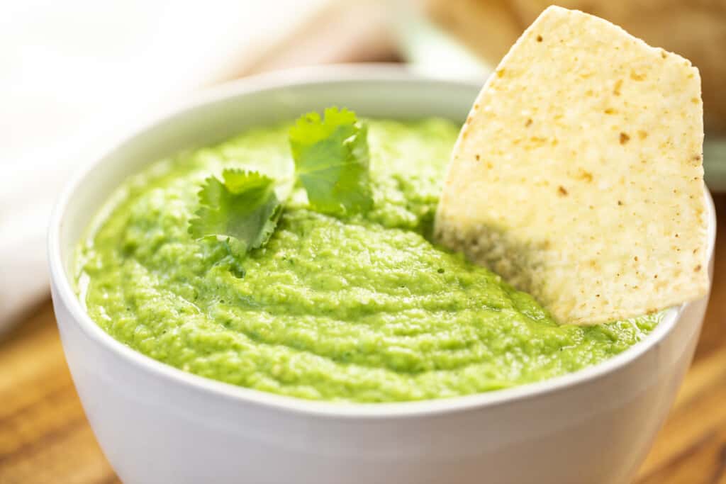 White bowl filled to the brim with tomatillo avocado salsa sits on a counter top with a tortilla chip dipped in.