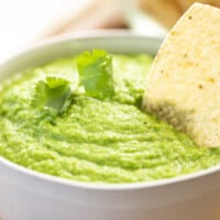 Creamy avocado tomatillo salsa sits in a bowl with tortilla chip dipped in.