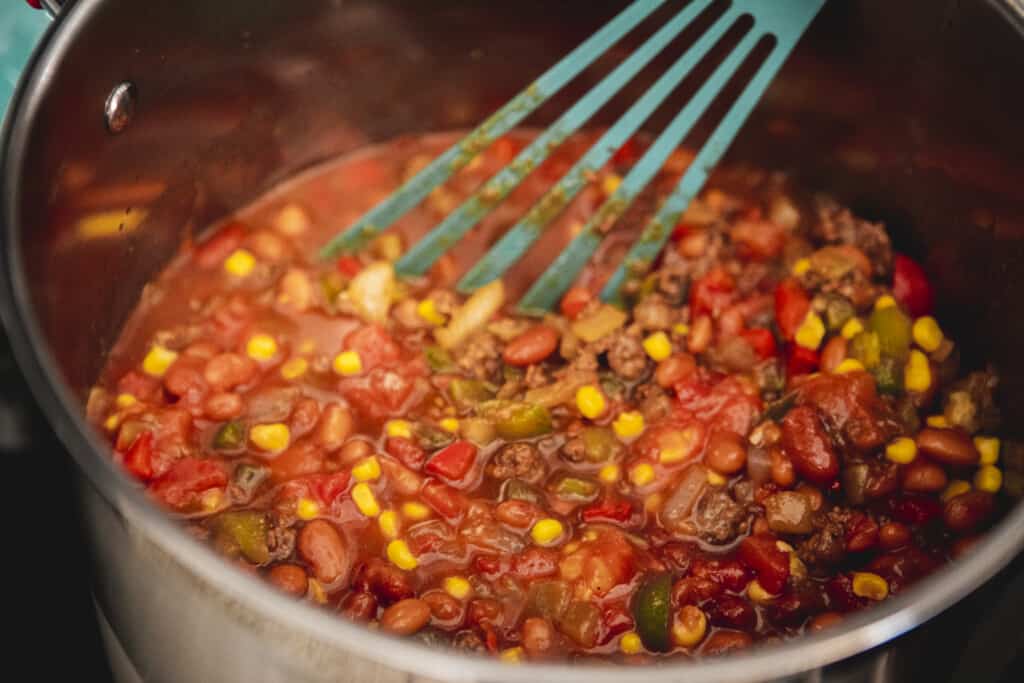 A spoon stirs chili in a large silver pot.