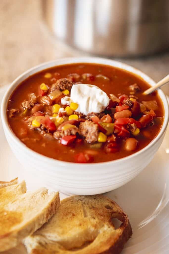 White ceramic bowl is filled with chili. A dollop of sour cream sits on top and a silver spoon sits inside the bowl. Toasted bread slices sit beside the bowl.