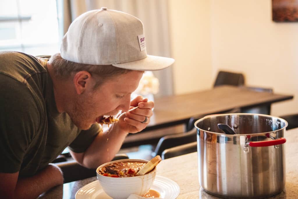 Dallin wearing a hat leans over a bowl of chili and takes a bite.