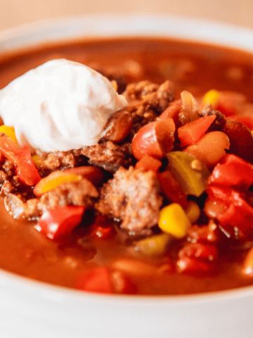 Bowl of chili sits on a white plate with dollop of sour cream on top.