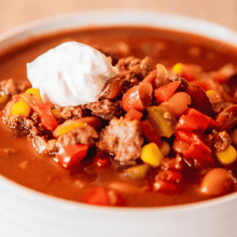 Bowl of chili sits on a white plate with dollop of sour cream on top.