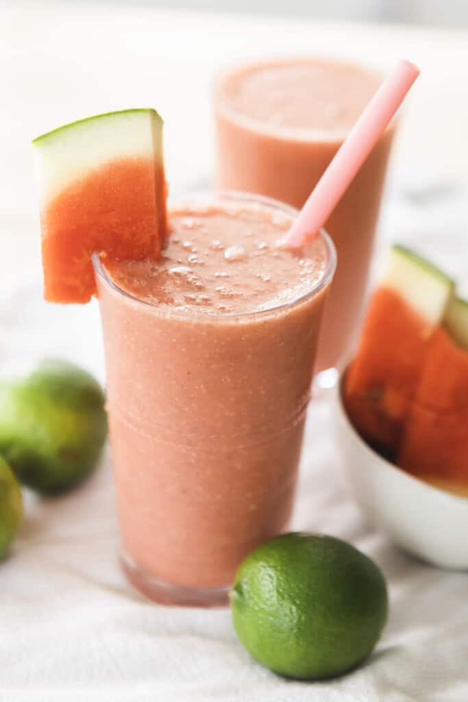 Glass of Watermelon Smoothie is sitting ready to enjoy. Wedge of watermelon hangs off the side with a pink silicone straw in glass.