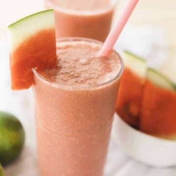 Glass of Watermelon Smoothie is sitting ready to enjoy. Wedge of watermelon hangs off the side with a pink silicone straw in glass.