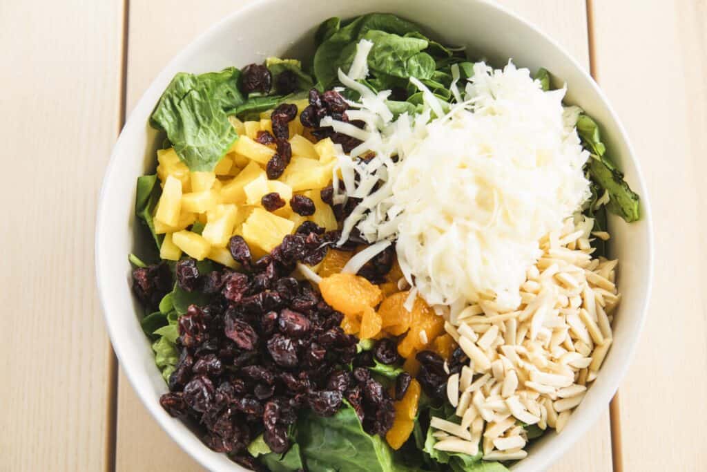 Pineapple, oranges, sliced almonds, swiss cheese, dried cranberries, and romaine sit in a large white bowl un-tossed.