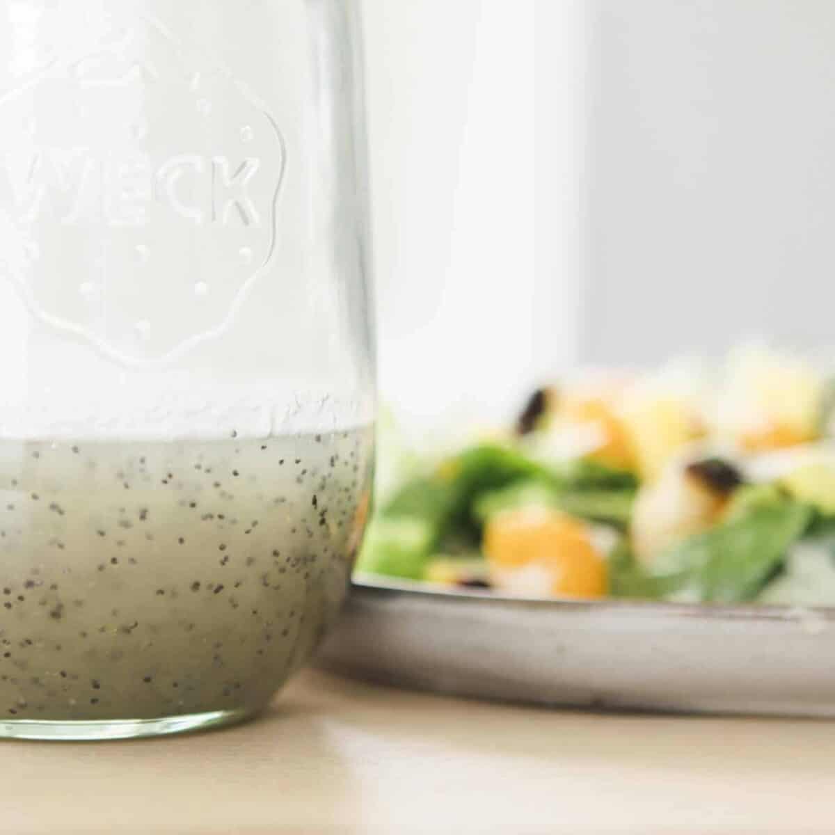 A bottle sits on the table top filled with homemade poppy seed dressing. Beside it sits a vibrant salad on a plate.