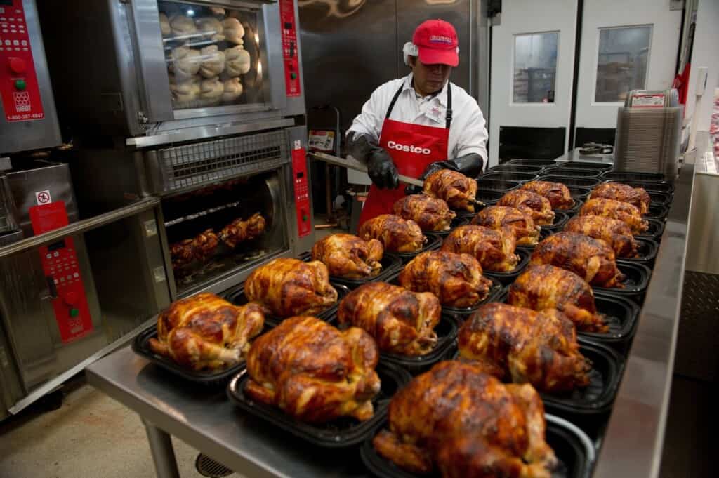 A Costco employee prepares a table full of rotisserie chickens by placing them in black containers.