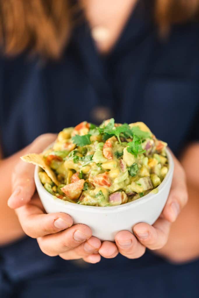Ashley stands holding out a bowl filled with fresh chunky mango avocado salsa. 