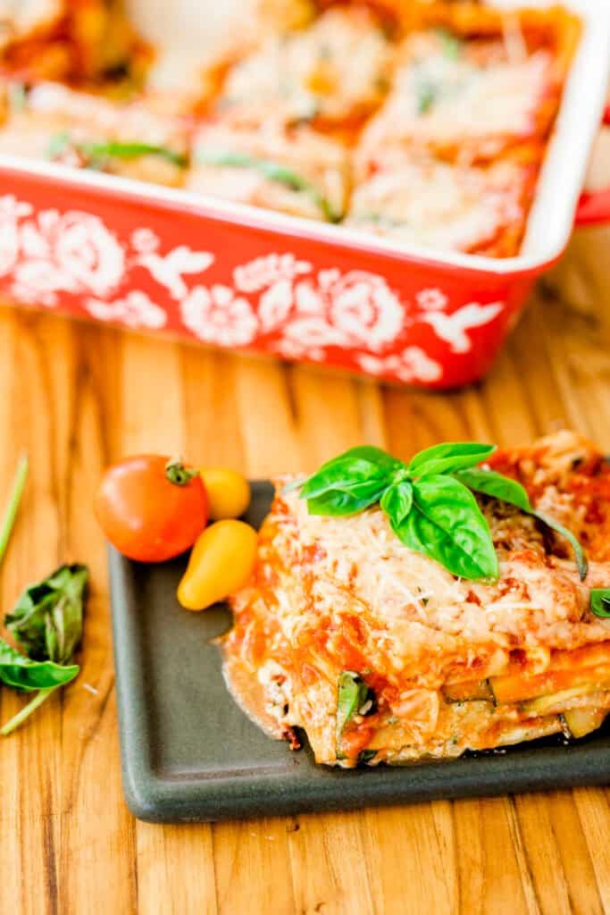 Slice of lasagna sits on a plate ready to eat. A casserole dish with slices of lasagna sits behind.