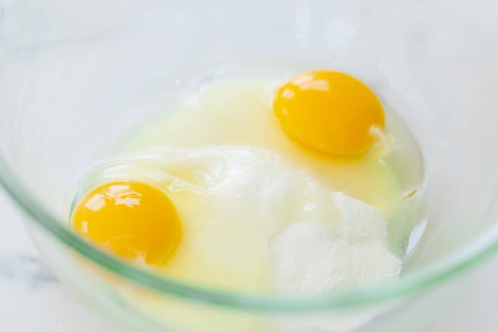 Two cracked eggs and sugar sit in a glass bowl ready to combine for a custard filling.