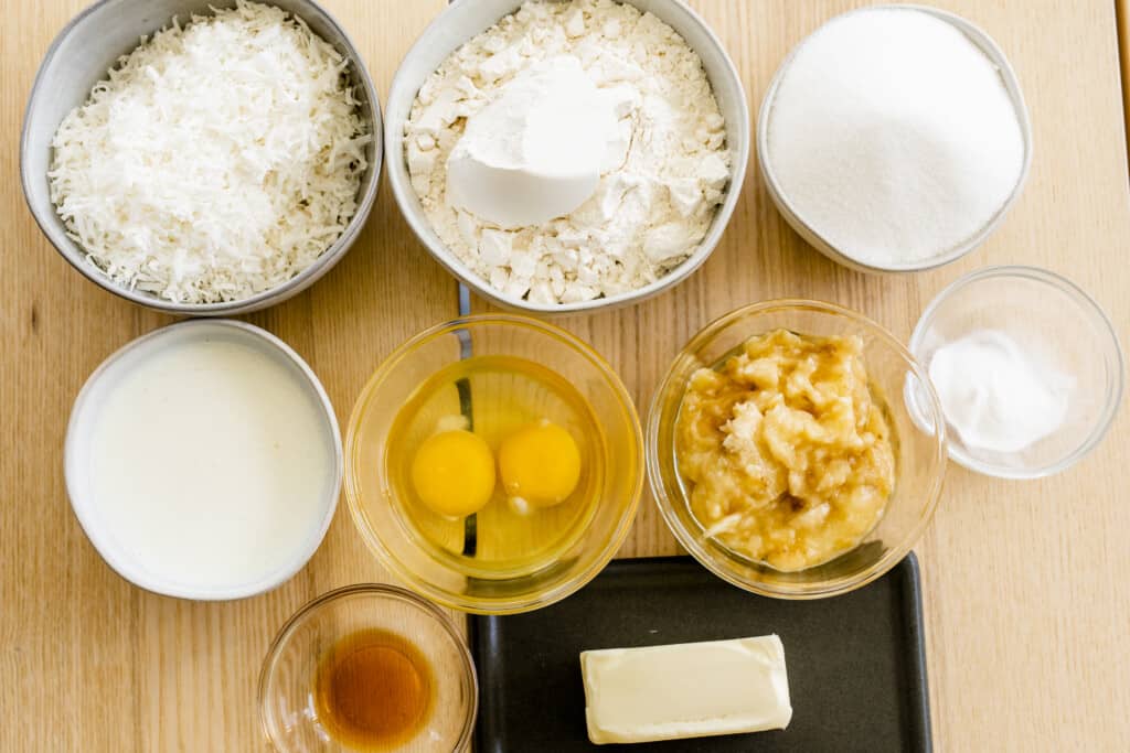 Ingredients for banana cake sit on the counter shredded coconut, flour, salt, sugar, soured milk, eggs, mashed bananas, vanilla and butter.