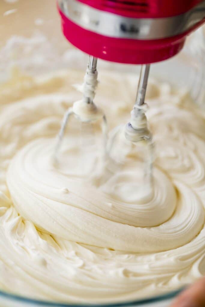 Coconut Cream Cheese Frosting is being whipped in a glass bowl by a hand mixer.