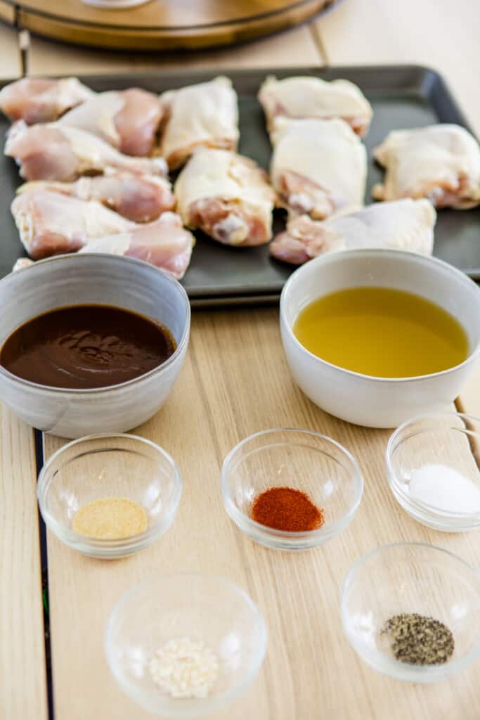 Raw drumsticks and thighs sit on a 13x9 cookie sheet with all the ingredients for barbecue chicken in foreground.