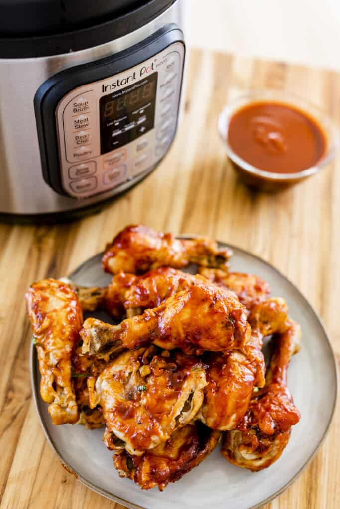 Chicken thighs and drumsticks sit on a plate stacked a top one another ready to eat. An Instant Pot cooker sits behind in the background next to a bowl of extra sauce.