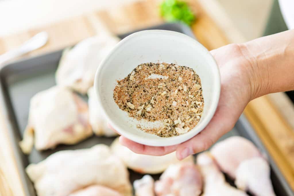 Homemade seasoning mix is combined in a bowl being held over a pan of uncooked chicken ready to be seasoned.