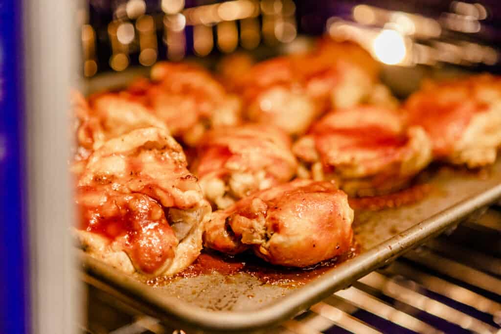 Barbecue chicken sits on a baking sheet in an oven on broil setting so that sauce caramelizes. 