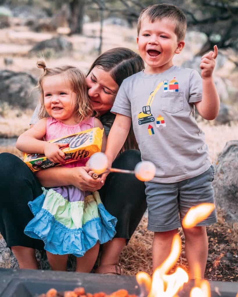 Ashley, James and George sit behind a campfire smiling and laughing while making smores.