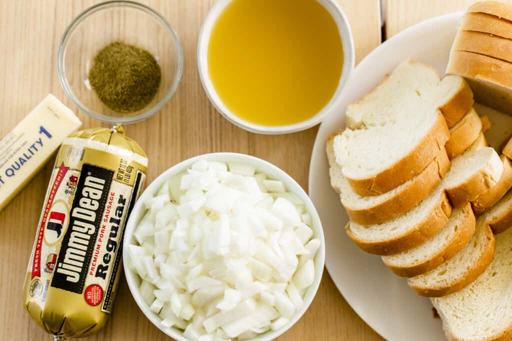 A loaf of bread sits on a plate, diced onions in a bowl, roll of sausage, a stick of butter, vegetable broth in a bowl and Italian seasonings sit on a countertop ready to be used in the recipe.