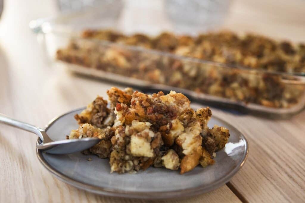 Serving of stuffing sits on plate with casserole dish behind.