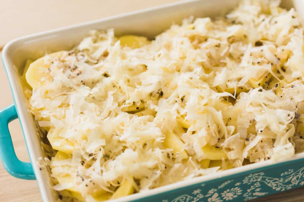 Slices of potato lie in a baking dish and are covered with shredded cheeses and topped with cream sauce for cheesy potatoes Au Gratin.