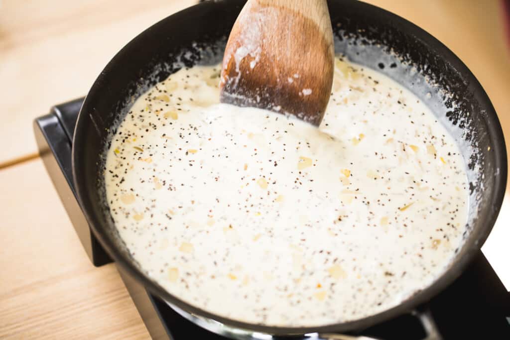 Cream sauce sits in a frying pan and is stirred with a wooden spoon ready to be poured over slices of potato for Au Gratin.