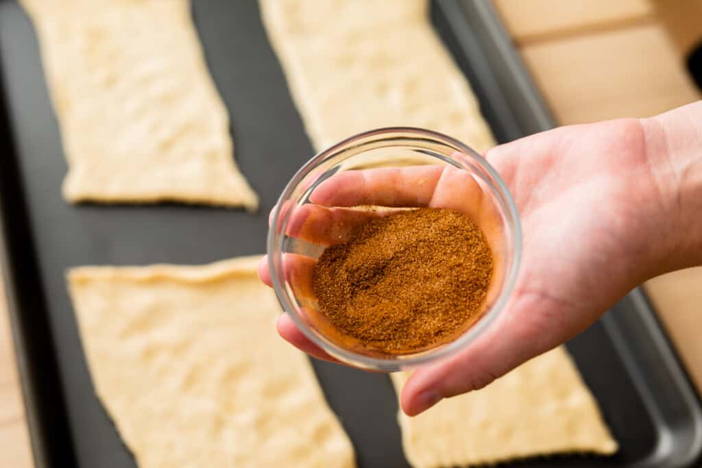 Crescent roll dough has been unrolled and separated into four rectangular sections. A hand is holding a small bowl of cinnamon sugar over the cookie sheet.