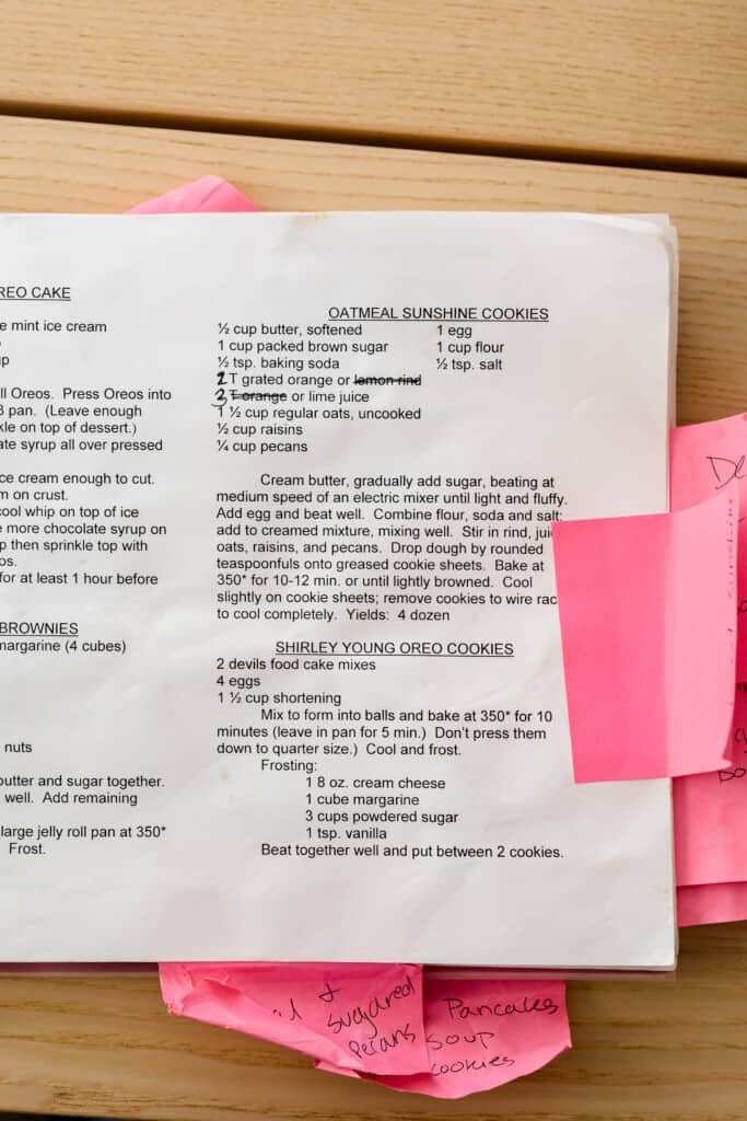 A worn and well loved cook book sits open on a table showing the recipe for Oatmeal Sunshine Cookies. Pink sticky notes with handwritten notes cover the edges of the pages.