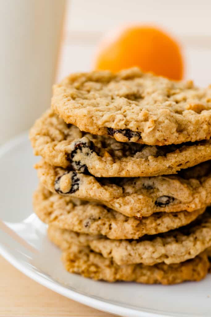 Six freshly baked cookies loaded with orange zest and raisins sit on top of each other on a white platter.