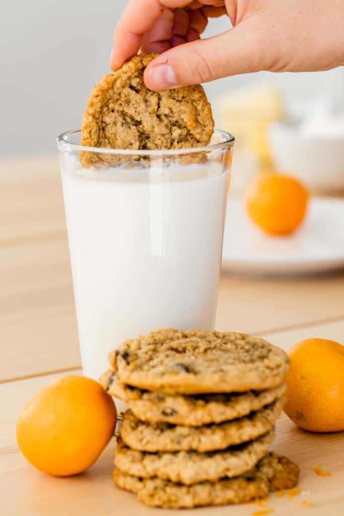 Ashley is holding a cookie in a tall glass of milk sitting behind a stack of cookies.