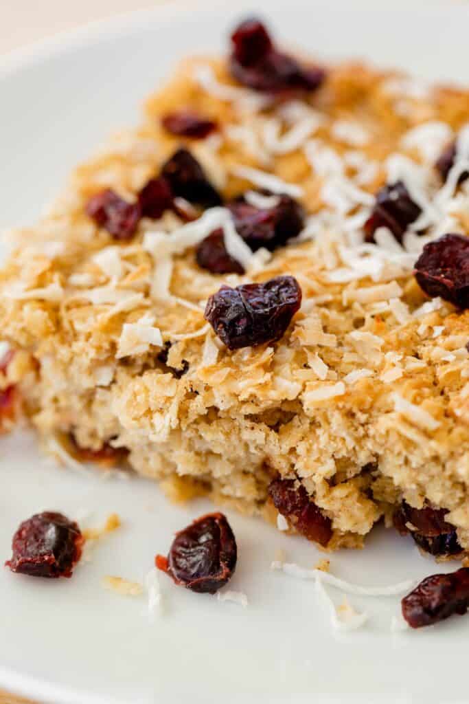 Baked Oatmeal breakfast bar sits on a plate with extra craisins and shredded coconut.