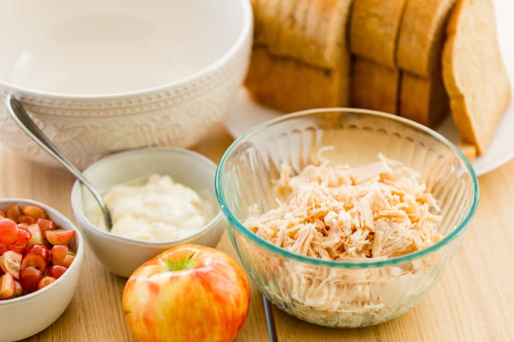 Ingredients for chicken salad are on a table top in separate bowls.