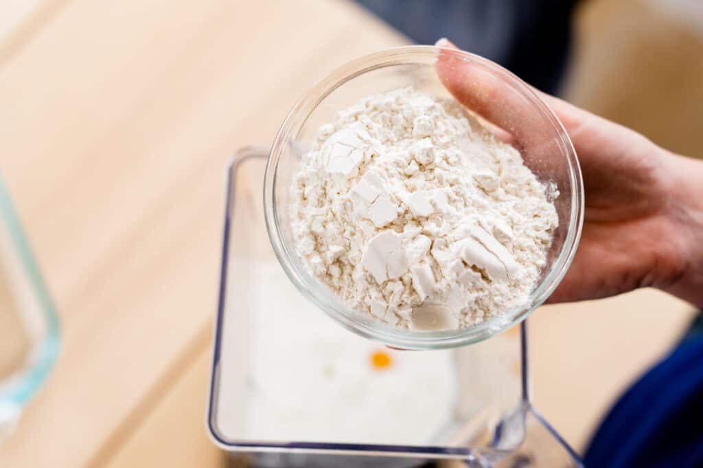 Ashley holds a small glass bowl of flour over the ingredient filled blender.