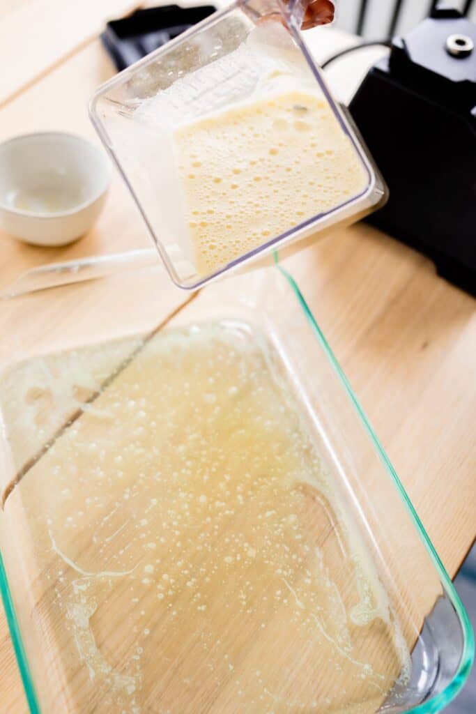 Glass casserole dish containing melted butter sits on the table. A blender filled with batter is held over the top ready to be poured into the dish.