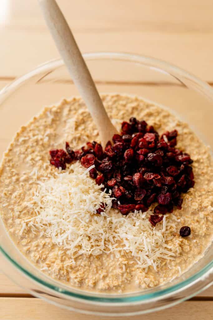 Glass bowl filled with oatmeal mixture has a layer of shredded coconut and craisins to be mixed in.