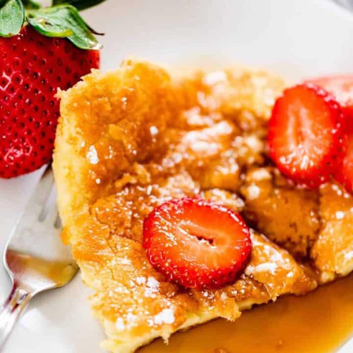 Slice of German Pancakes sit on a small round plate. Pancake is covered in caramel brown syrup and topped with sliced strawberries.