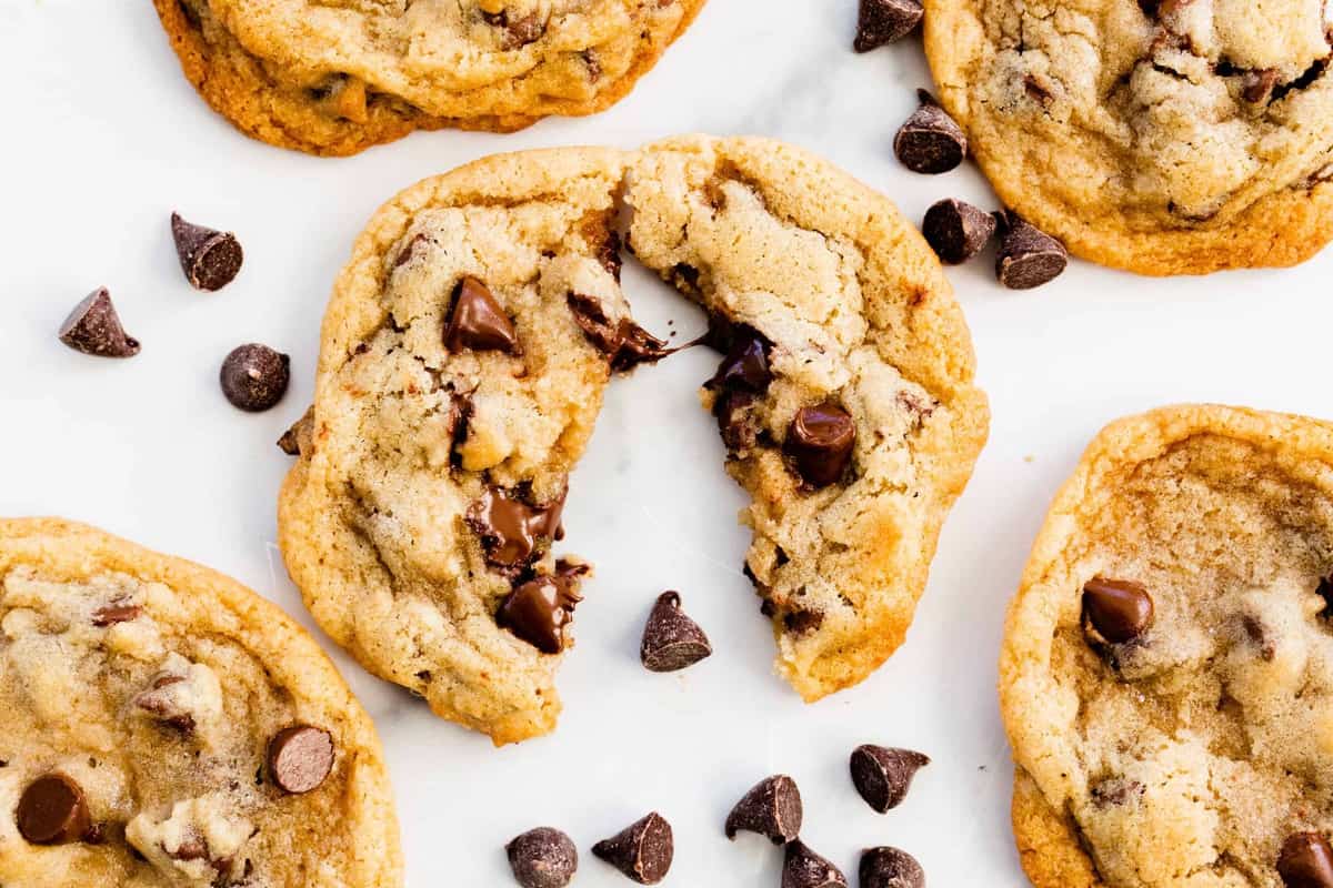 A big fresh, chocolate chip cookie sits broken in half on the counter top surrounded by other cookies and scattered chocolate chips.