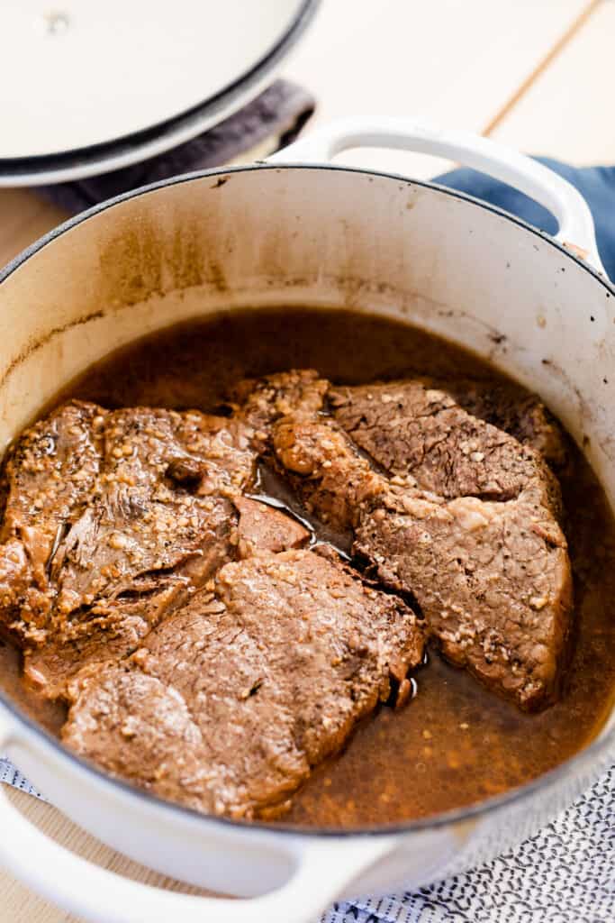 A cast iron pot sits on the stove with a perfectly cooked chuck roast sitting in au jus.