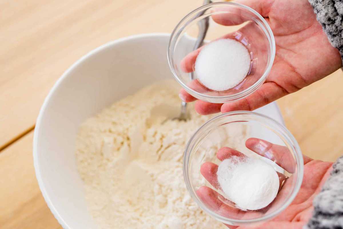 Ashley holds two small bowls with salt and baking soda over a bowl of flour.