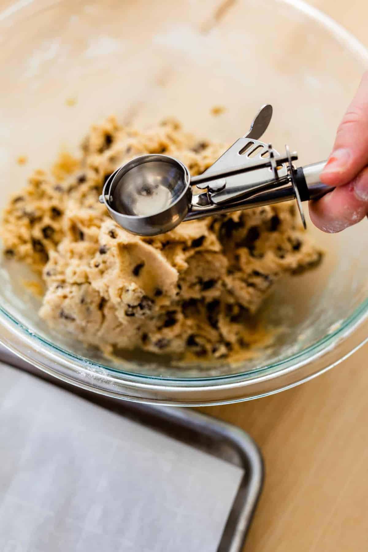 Ashley holds a cookie scoop over a bowl of cookie dough next to a baking sheet.