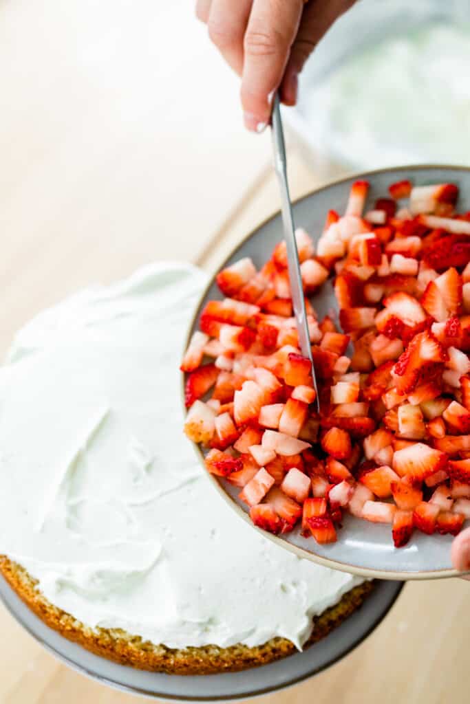 A bowl of freshly diced strawberries is held over the strawberry cake.