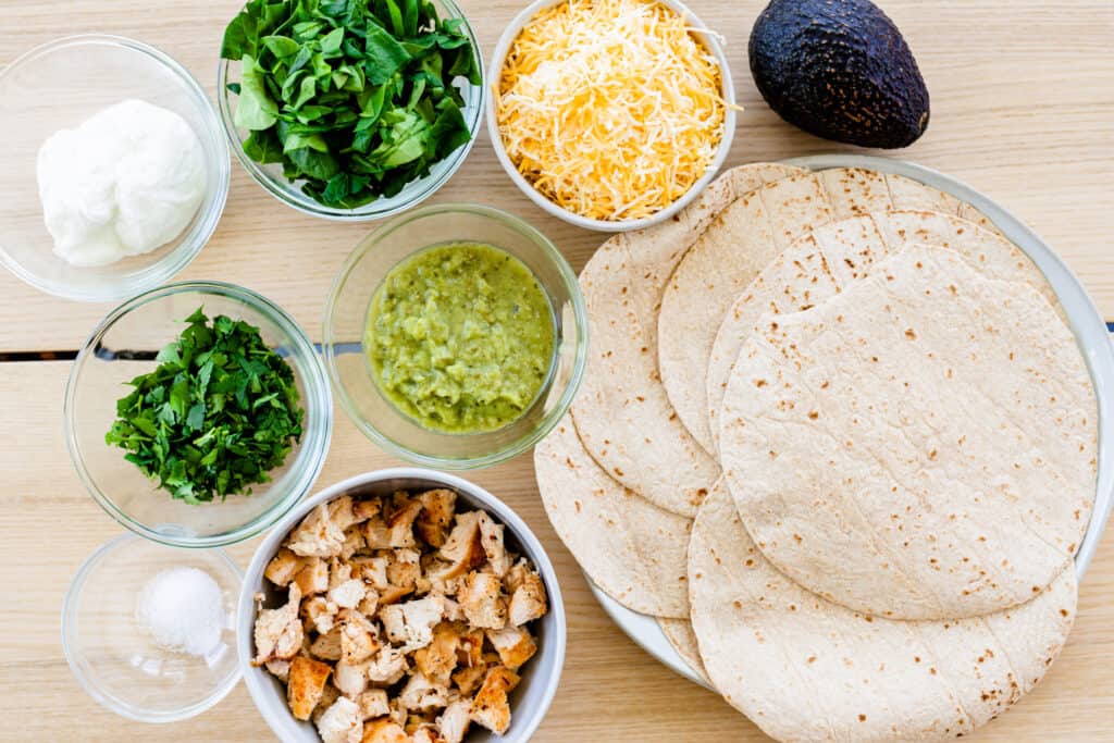 Ingredients for green machine quesadilla sit on the counter top in small glass and ceramic bowls. Cilantro, spinach, sour cream, salsa verde, grilled chicken, avocado, salt, cheese and tortillas.