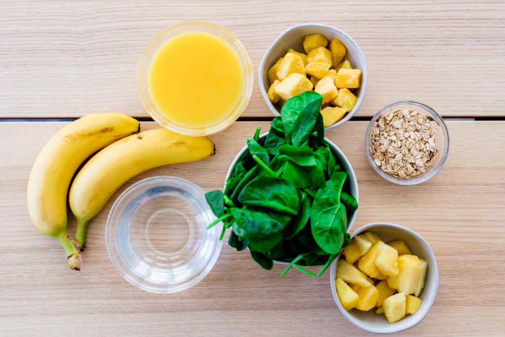 Ingredients for smoothie sit on table in small glass bowls. Banana, ice, juice, pineapple, spinach, oatmeal, mango.