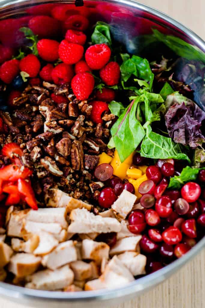 Candied walnuts have been added to a large metal bowl sits on the countertop filled with mixed greens, grilled chicken, vibrant berries and mango.