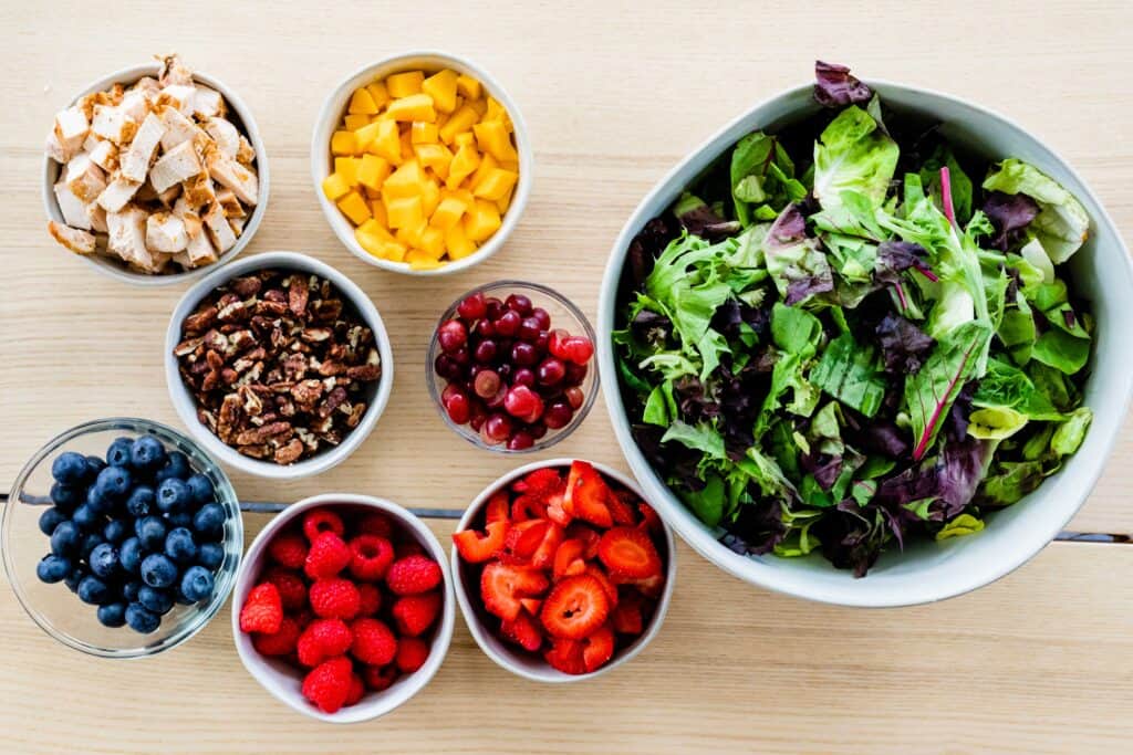 Blueberries, Raspberries, Strawberries, Grapes, Mangoes, Mixed Greens, Grilled Chicken sit in individual bowls on a table top ready to be combined.