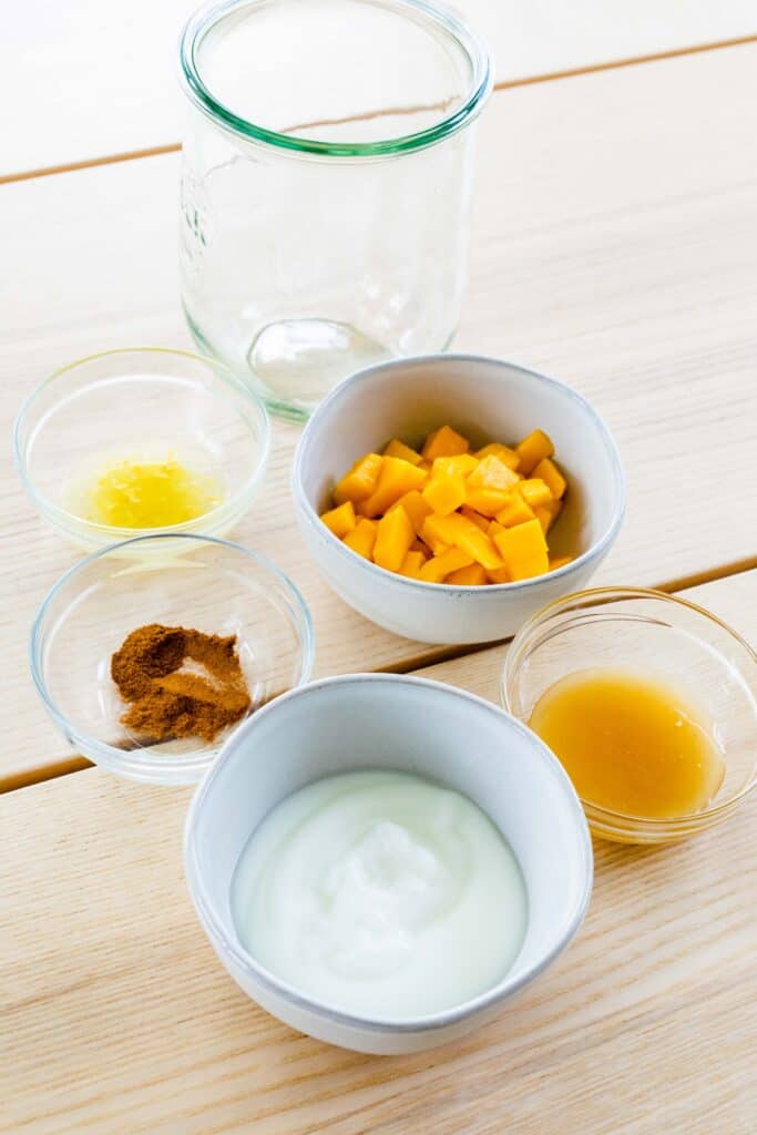 Mangoes, yogurt, lemon juice and zest, honey sit in small bowls on a table top in small individual bowls.