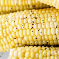 Cooked corn on the cob sits stacked on a white plate sprinkled with pepper, ready to eat.