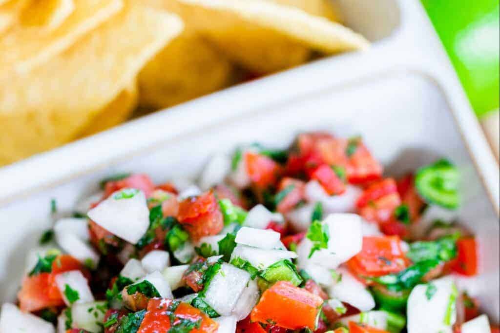 Chilled Pico de Gallo sits in a white ceramic serving dish alongside crispy corn tortilla chips.